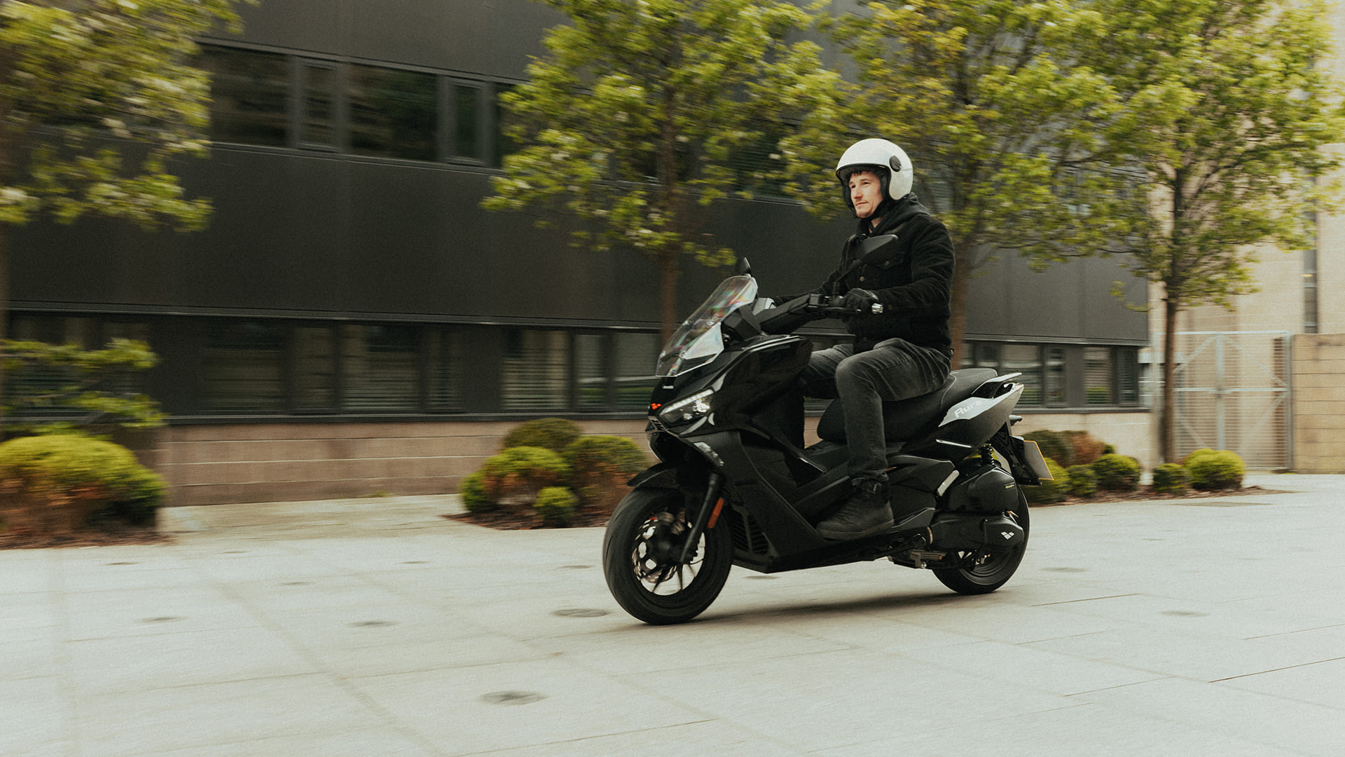 Rider on Aura 125 cruising past modern office with greenery